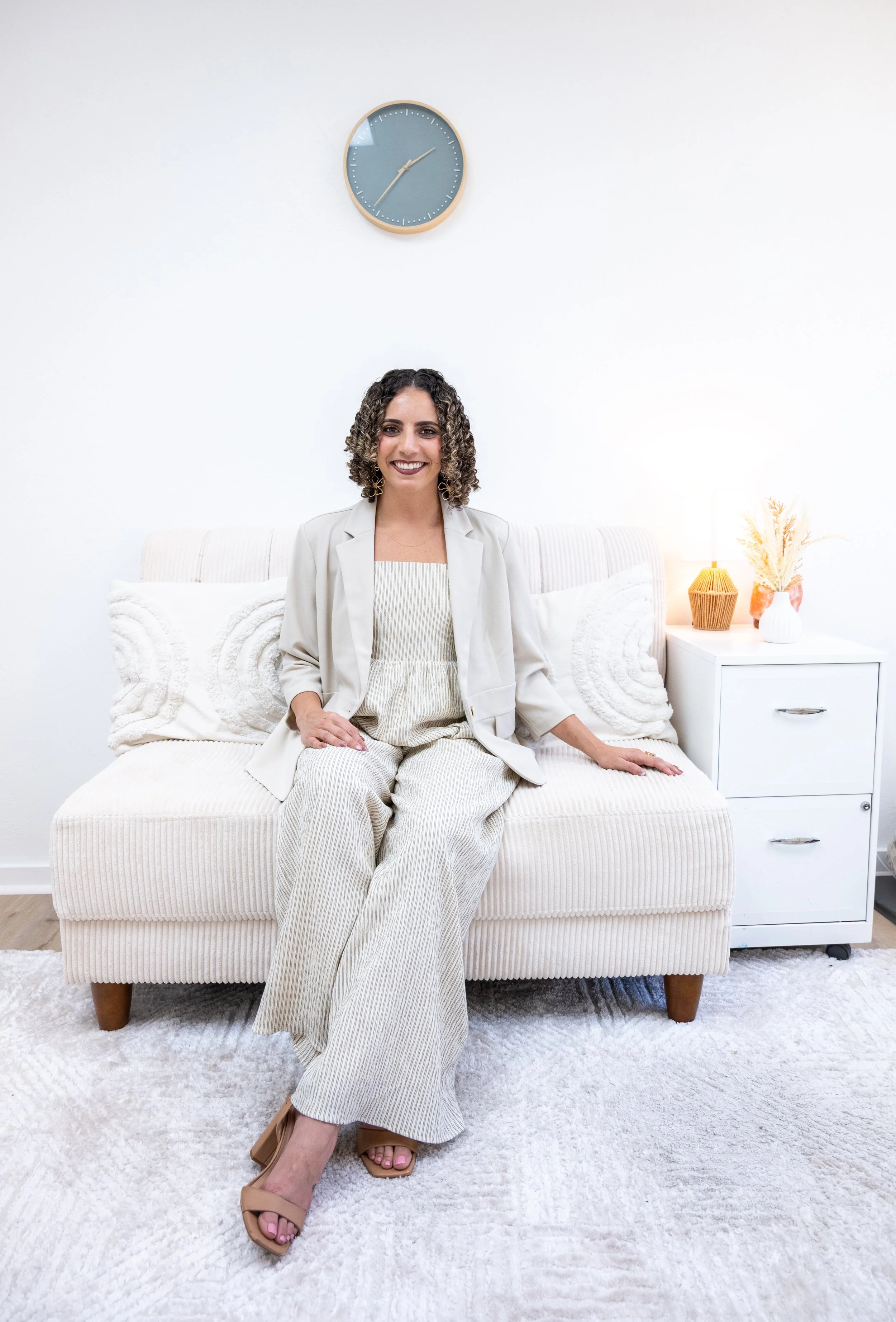A woman with curly hair sits on a white sofa in a bright, minimalistic room. She is smiling, wearing a light-colored blazer and matching loose pants, with tan open-toe heels. Behind her is a white wall with a blue clock above her head, and a white side table with a lamp and decorative plants on her right.