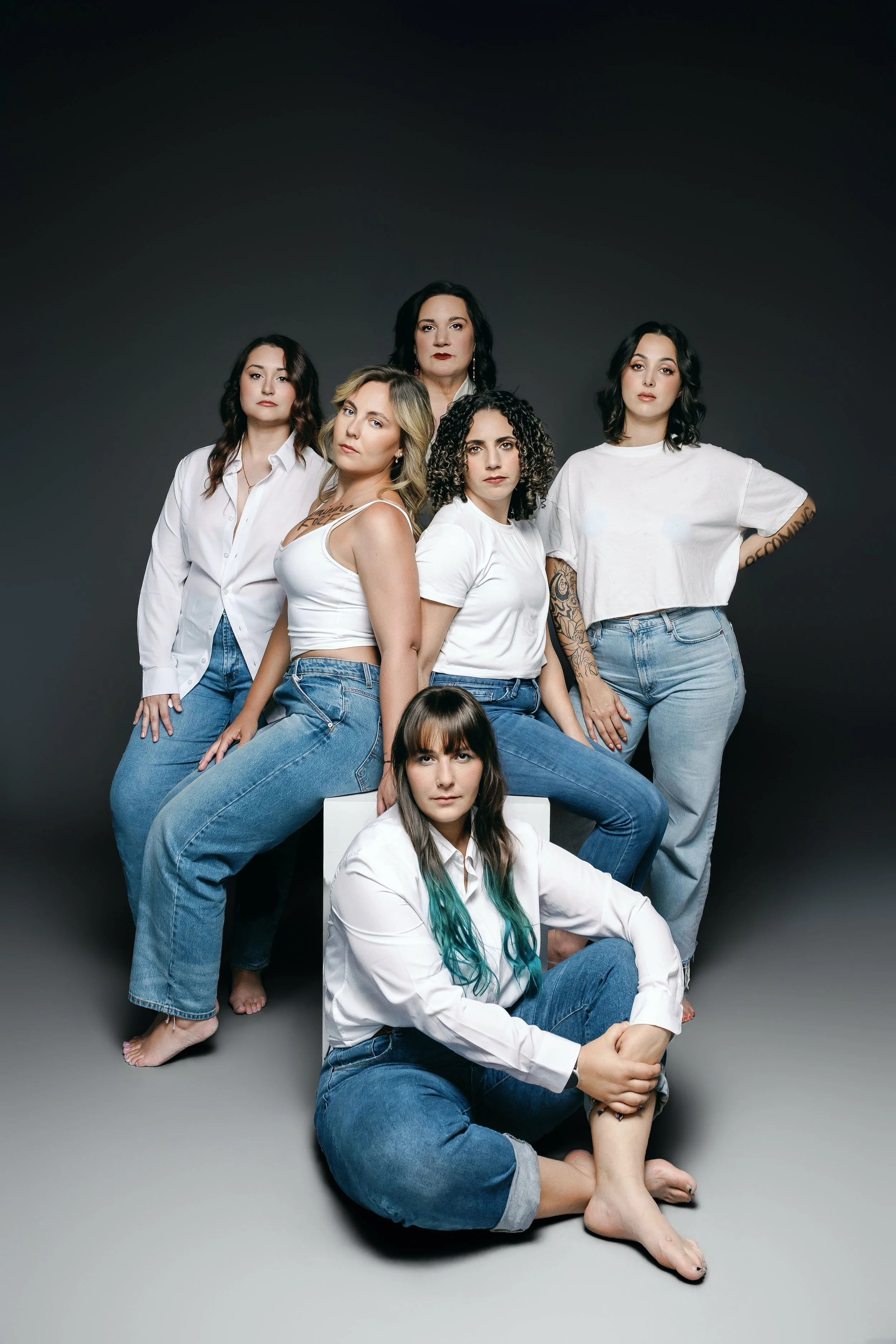 A group of eight female sexual assault survivors with varying hair colors against a dark background. They are wearing casual clothing, primarily white tops and jeans. Some are standing, some sitting or kneeling, with serious or neutral expressions.