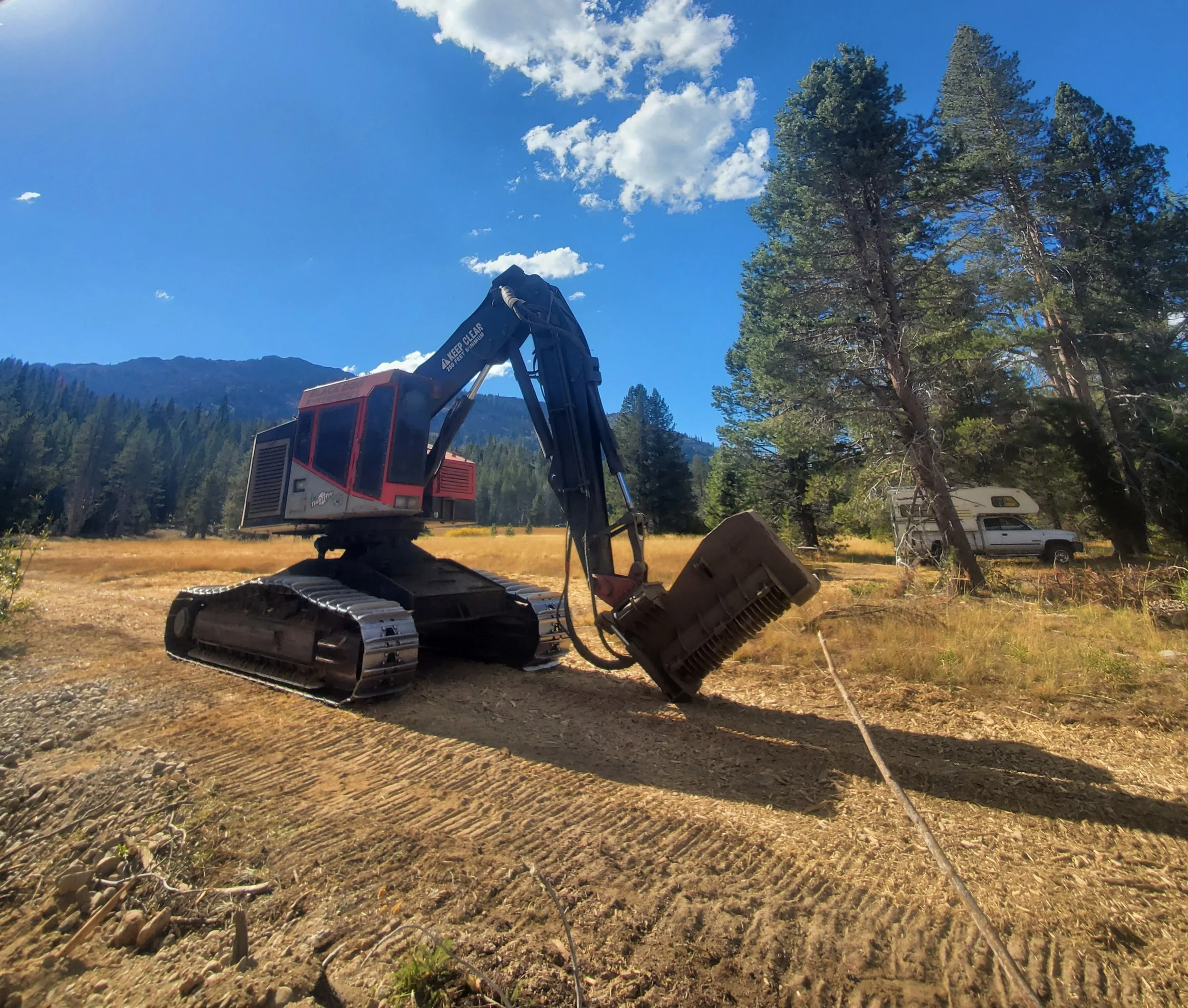 A construction excavator working on a plot of land with trees and mountains in the background, under a bright blue sky with white clouds.