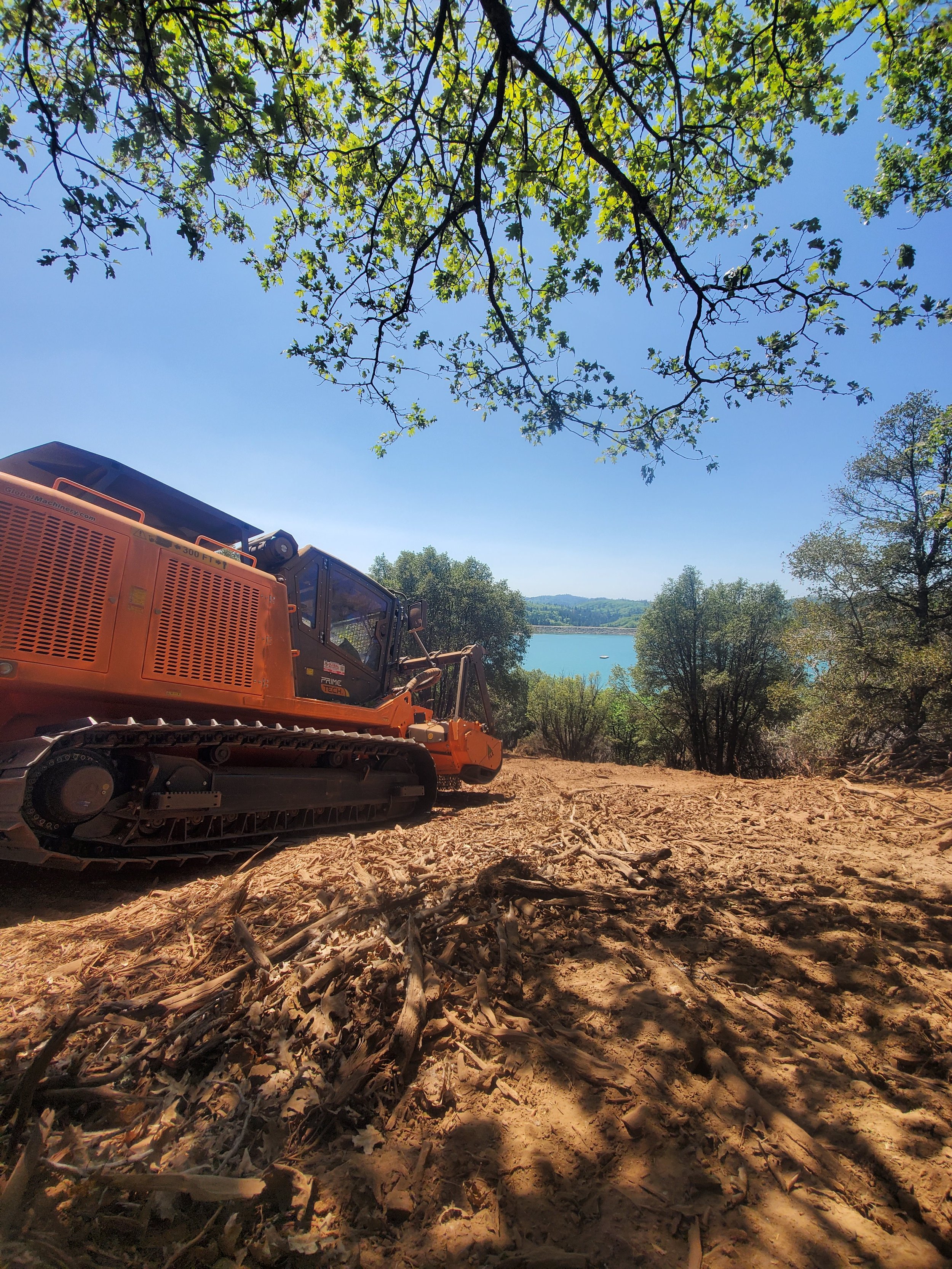 A construction or logging site with a large orange excavator on a dirt ground surrounded by trees, with a lake and hills visible in the background under a clear blue sky.