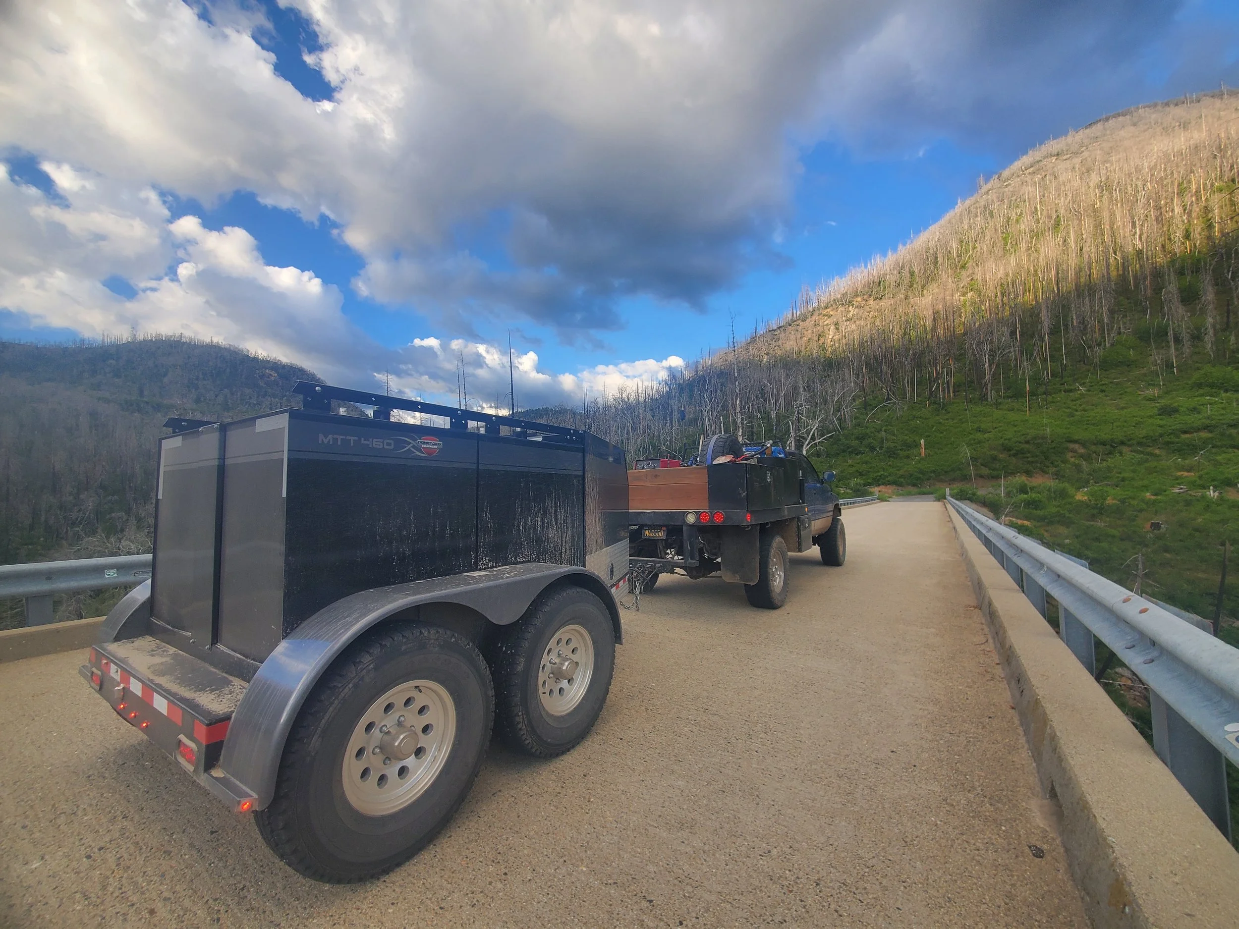 A flatbed truck parked on a mountain road with a landscape of burnt trees on the hillside and a partly cloudy sky.