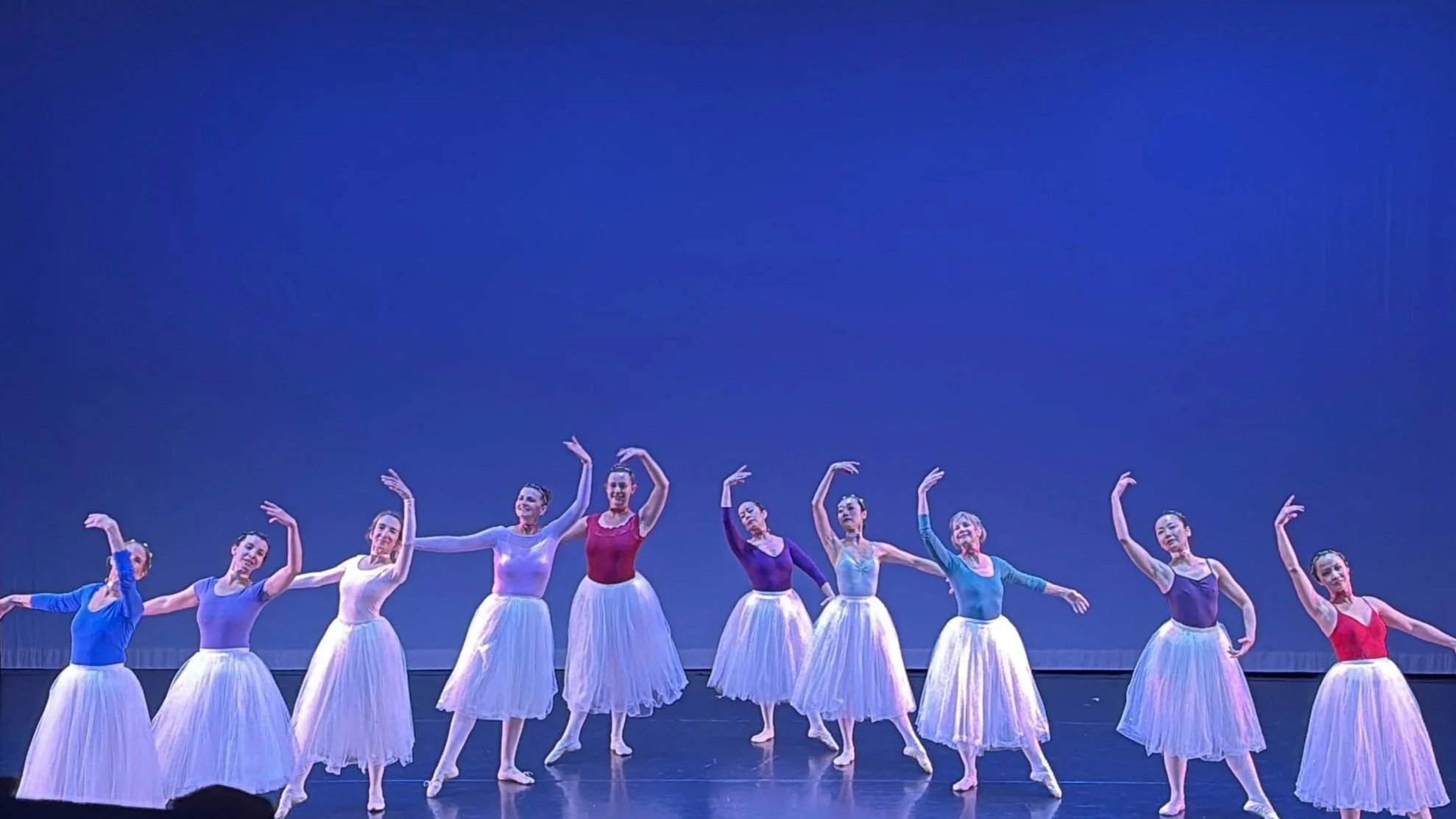 A group of ballet dancers performing on stage, dressed in colorful tops and white skirts, striking various elegant poses against a blue backdrop.