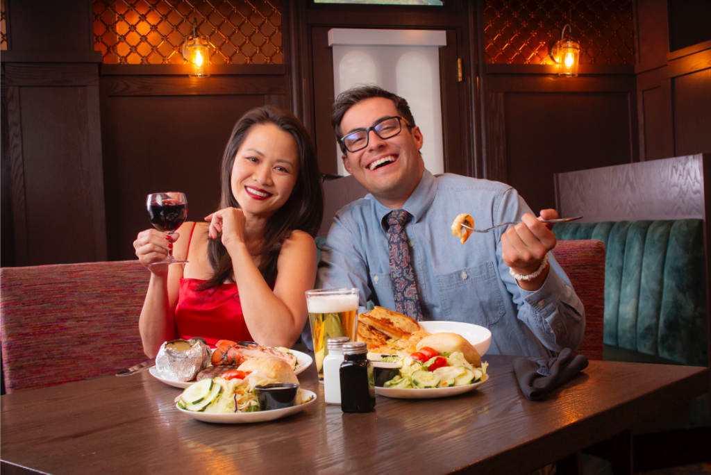 A smiling man and woman are sitting at a restaurant table with plates of food and drinks, enjoying a meal together.