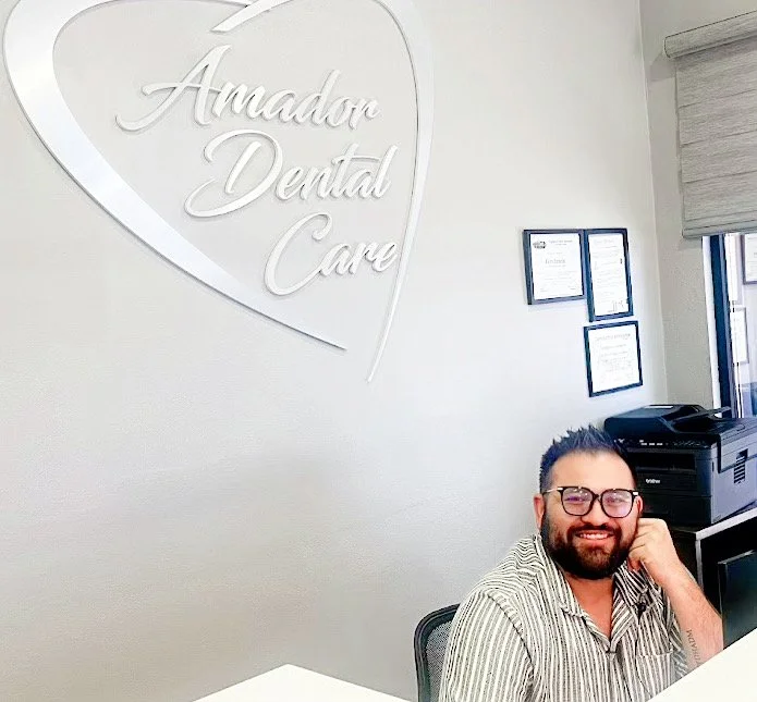 A smiling man with glasses sits at a reception desk in a dental office with a sign reading 'Amador Dental Care' on the wall behind him. Certificates and a printer are visible in the background.