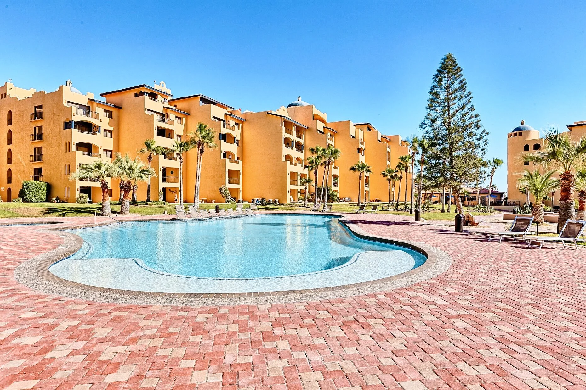 Outdoor swimming pool surrounded by lounge chairs, palm trees, and a large orange apartment complex, clear sky.
