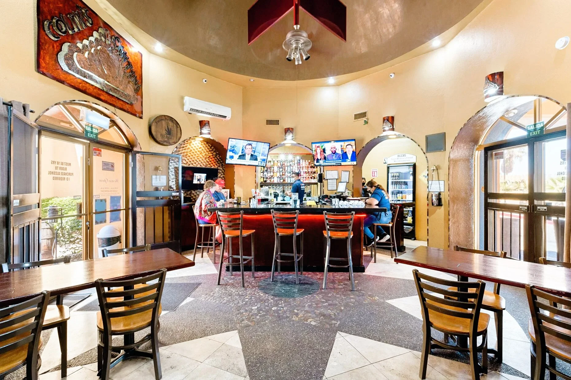 Interior of a bar with a circular bar counter, chairs, wall-mounted TVs, and tables. The room has arched entrances and ceiling lights.