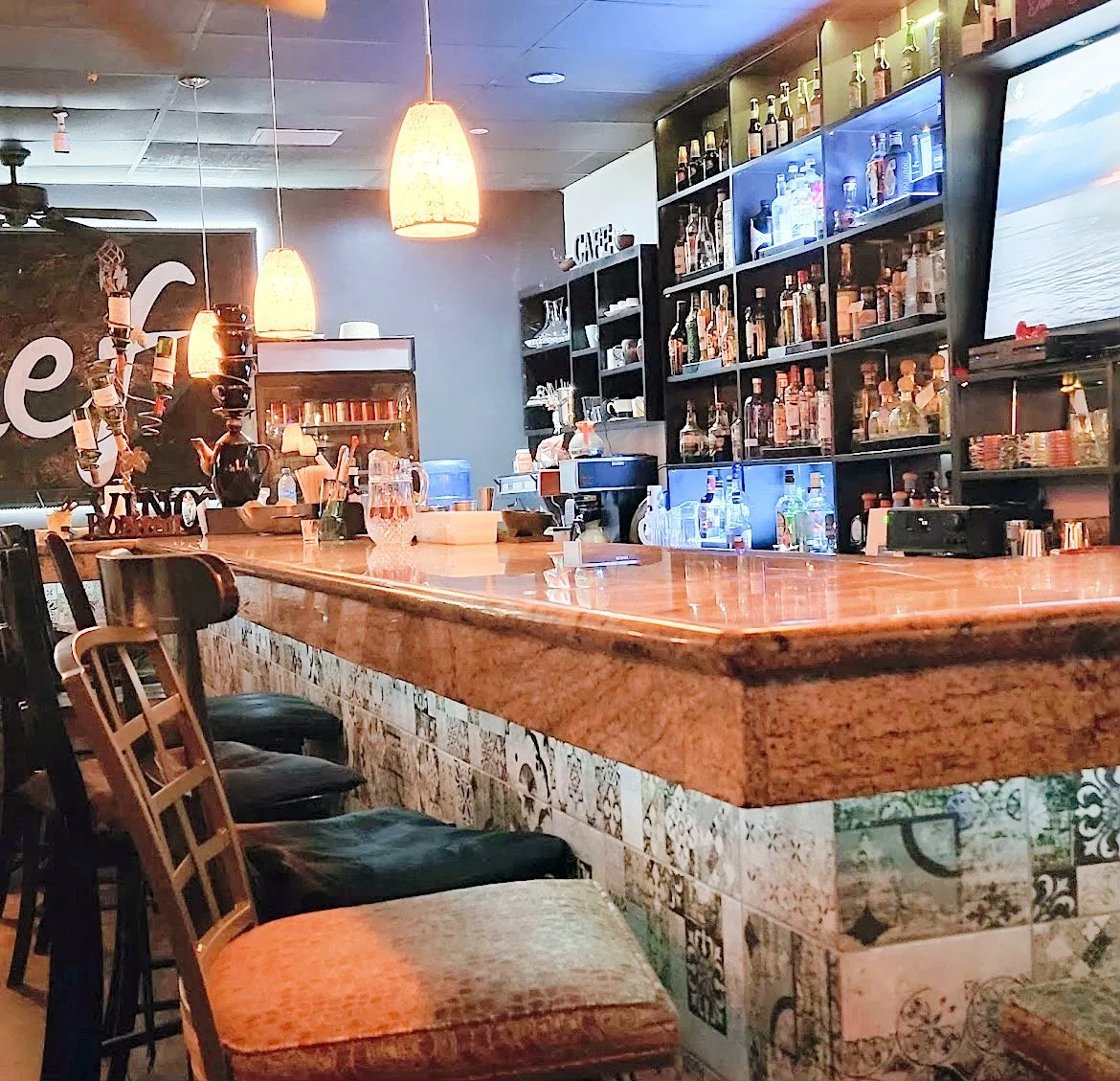 A cozy bar interior with a wooden countertop, upholstered bar stools, hanging pendant lights, and shelves stocked with various liquor bottles.