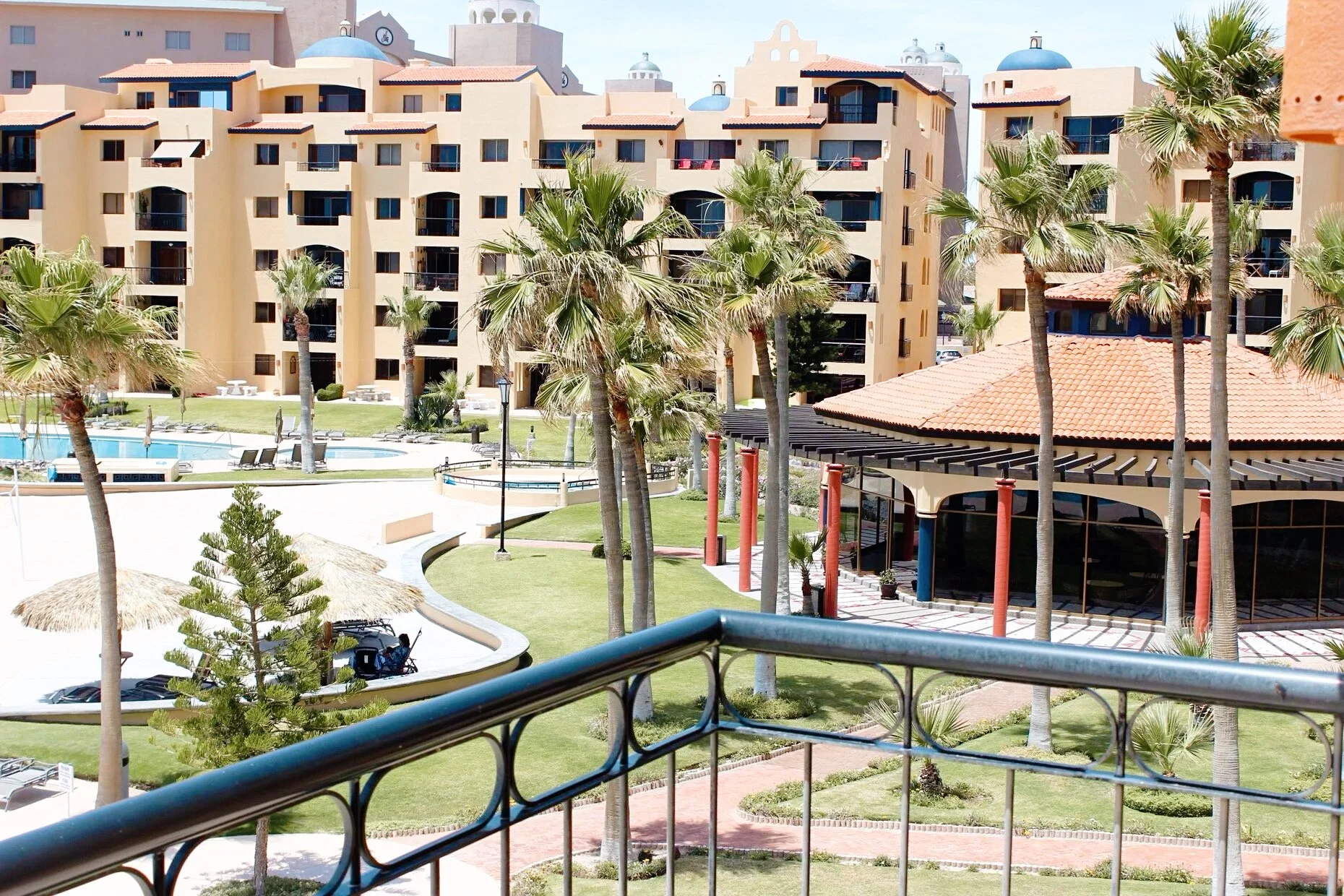 Resort complex with palm trees, swimming pool, and sandy pathways, surrounded by tan buildings with terracotta roofs.