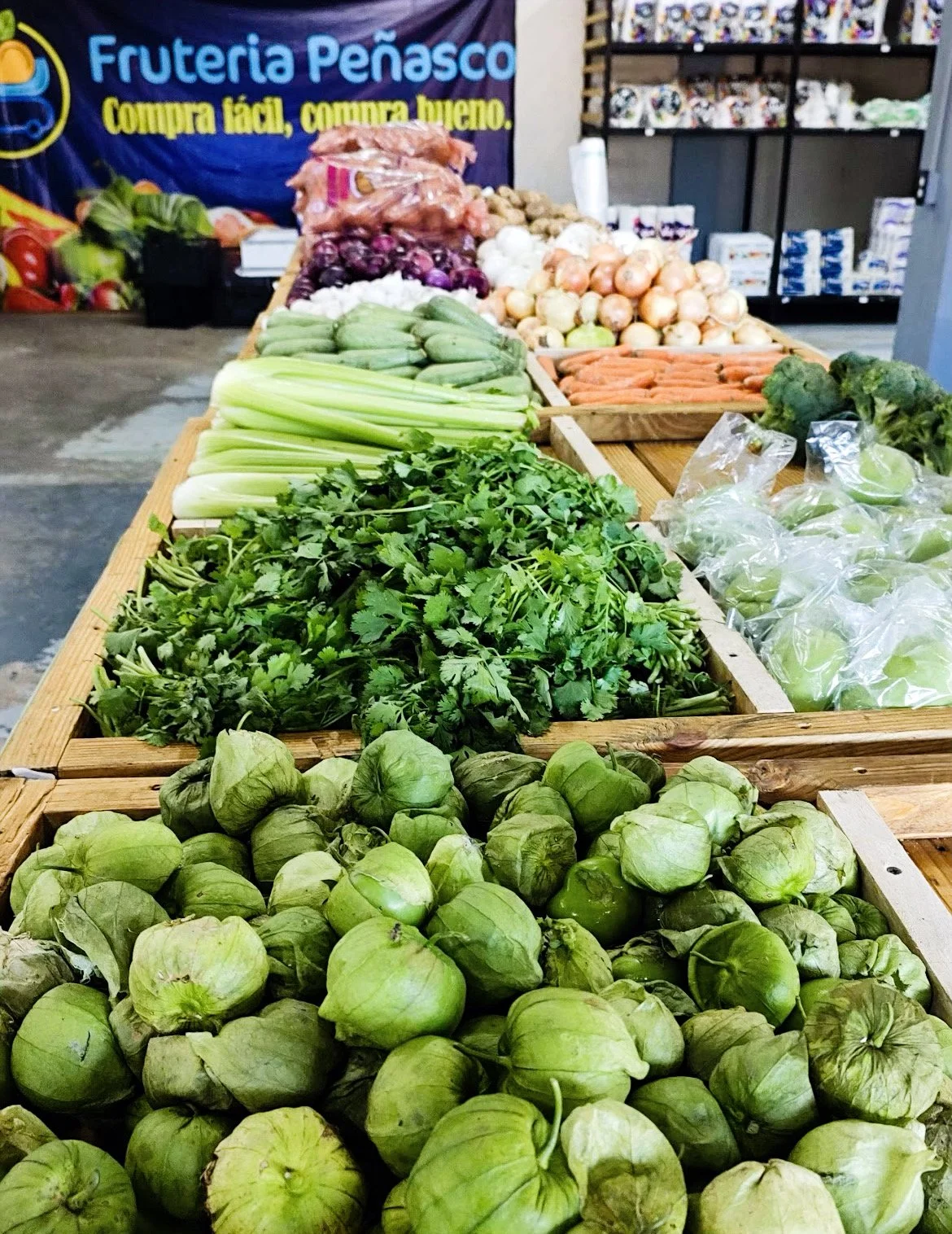 Display of fresh vegetables at a market, including tomatillos, cilantro, celery, zucchini, purple onions, white onions, carrots, and potatoes. Sign reads 'Fruteria Peñasco' in the background.
