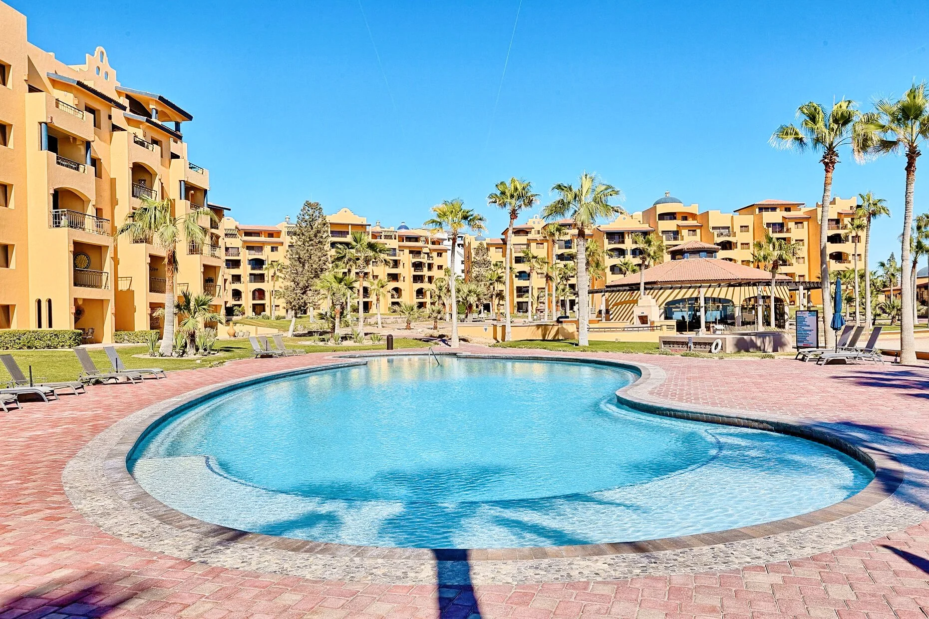 Resort with a large swimming pool surrounded by palm trees and tan-colored buildings under a clear blue sky.