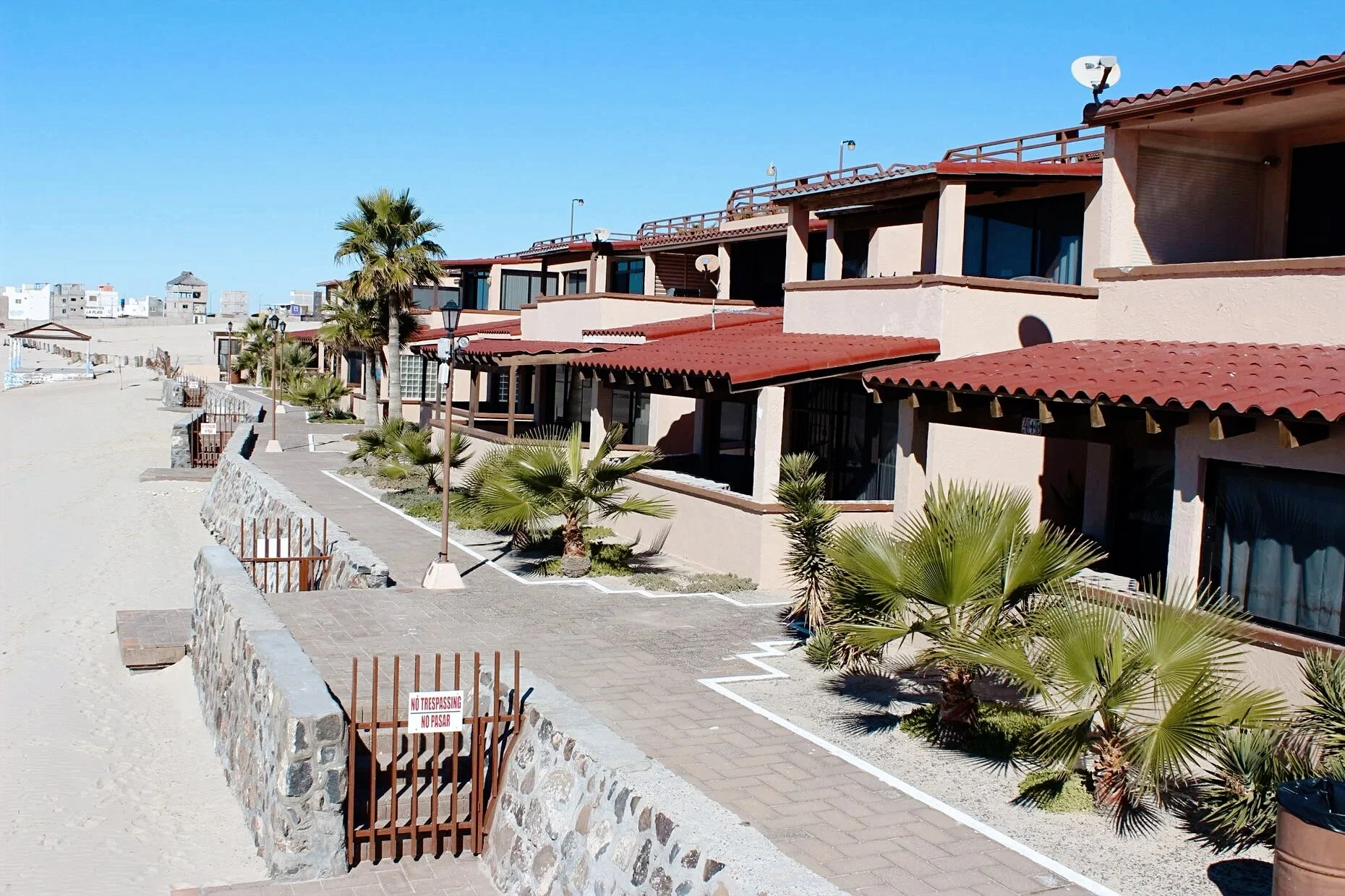 Beachfront row of houses with red roofs, palm trees, and a stone walkway adjacent to a sandy beach.
