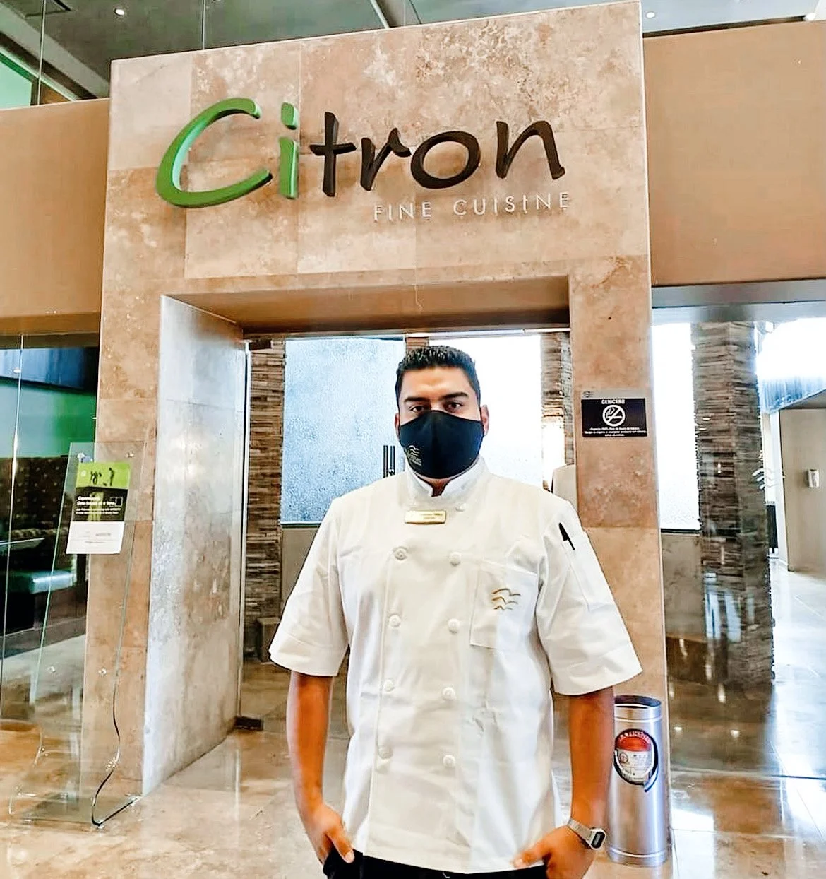Chef in white uniform and black mask standing in front of Citron Fine Cuisine entrance.