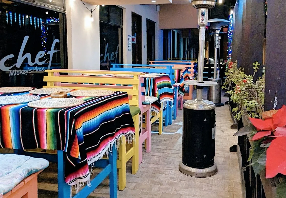 Outdoor restaurant seating area with colorful tables and benches, covered in striped blankets, and surrounded by patio heaters and plants.