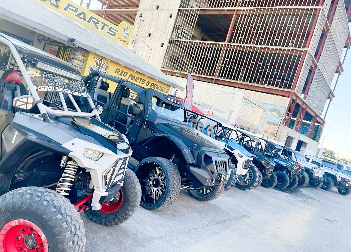 Lineup of off-road vehicles, including ATVs and side-by-sides, outside a rental shop with a sign for parts and rentals. A multi-story building is in the background.
