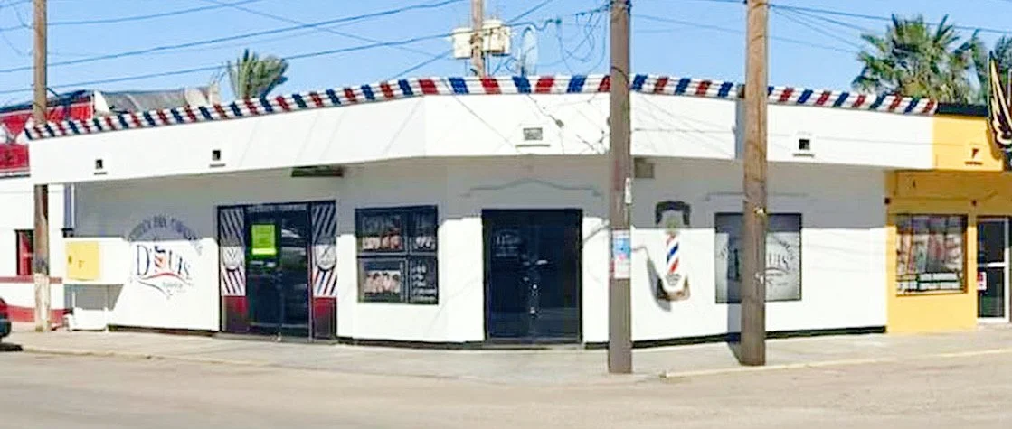 Exterior of a barbershop with a red, white, and blue striped roof trim. There are barbershop signs on the building, and palm trees are visible in the background.