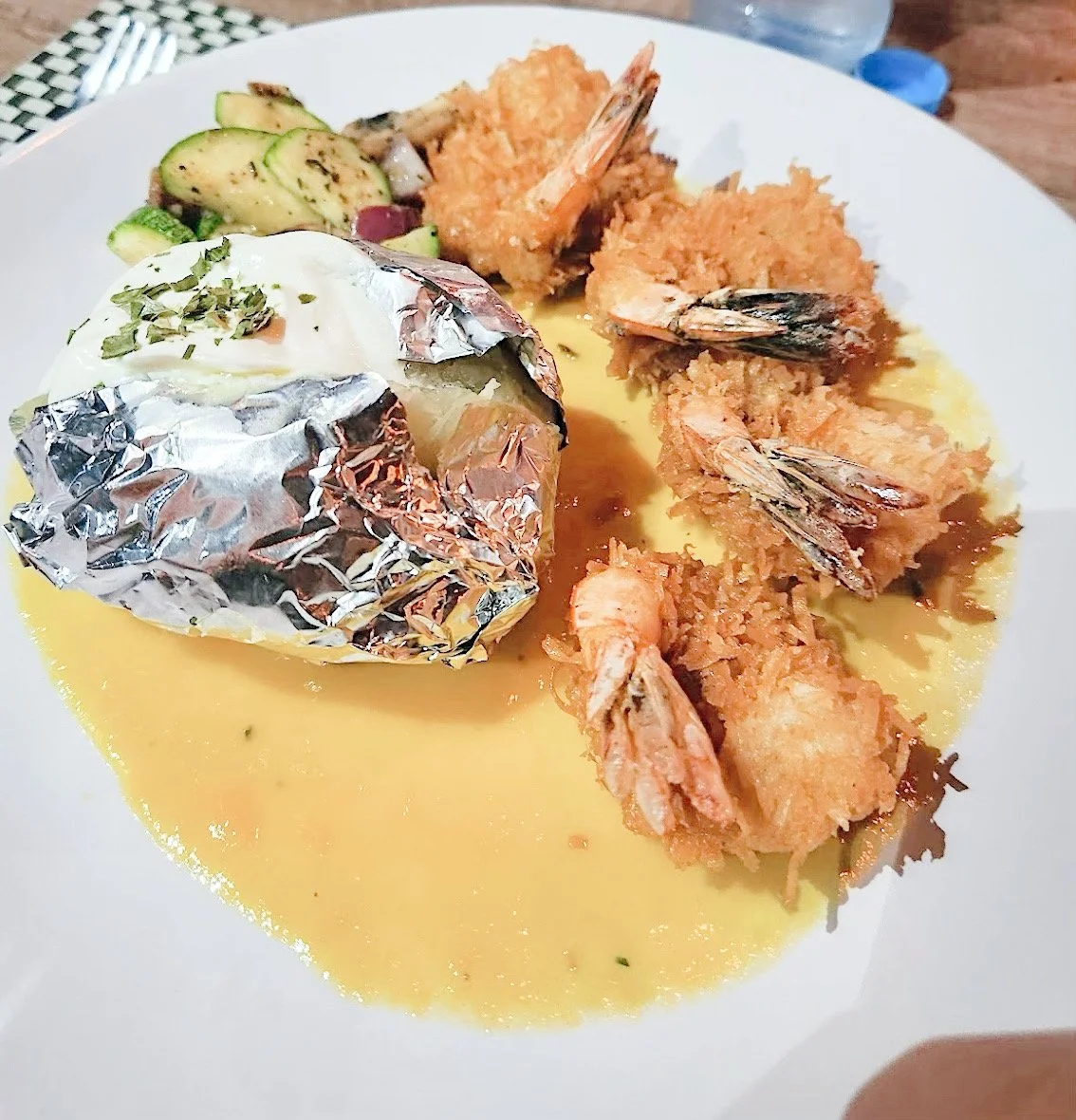 Coconut shrimp with yellow sauce, foil-wrapped baked potato topped with sour cream, and sautéed vegetables on a white plate.
