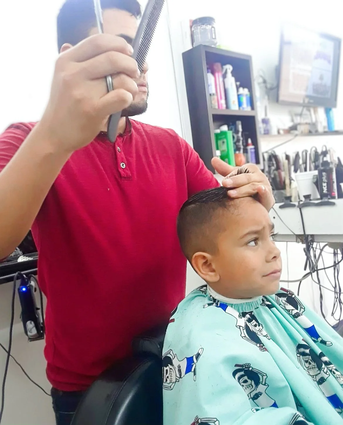 Barber cutting a young boy's hair in a salon, using scissors and a comb. The boy is seated and wearing a cape with cartoon characters. Products and tools are visible in the background.
