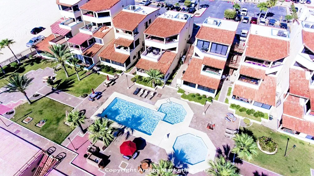 Aerial view of a residential complex with red-tiled roofs, swimming pools, and landscaped gardens with palm trees.