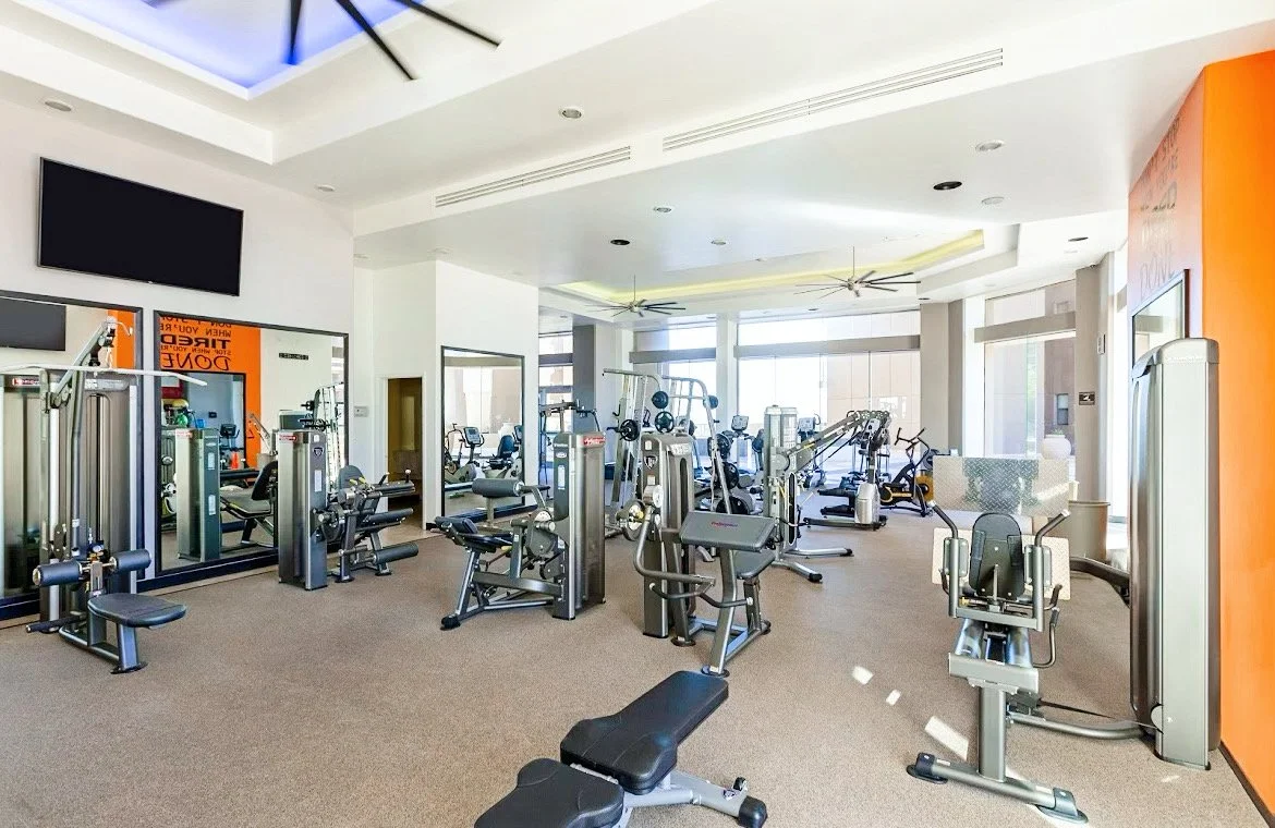 Modern gym with exercise machines and equipment, including treadmills, weight machines, and stationary bikes, inside a well-lit room with large windows.