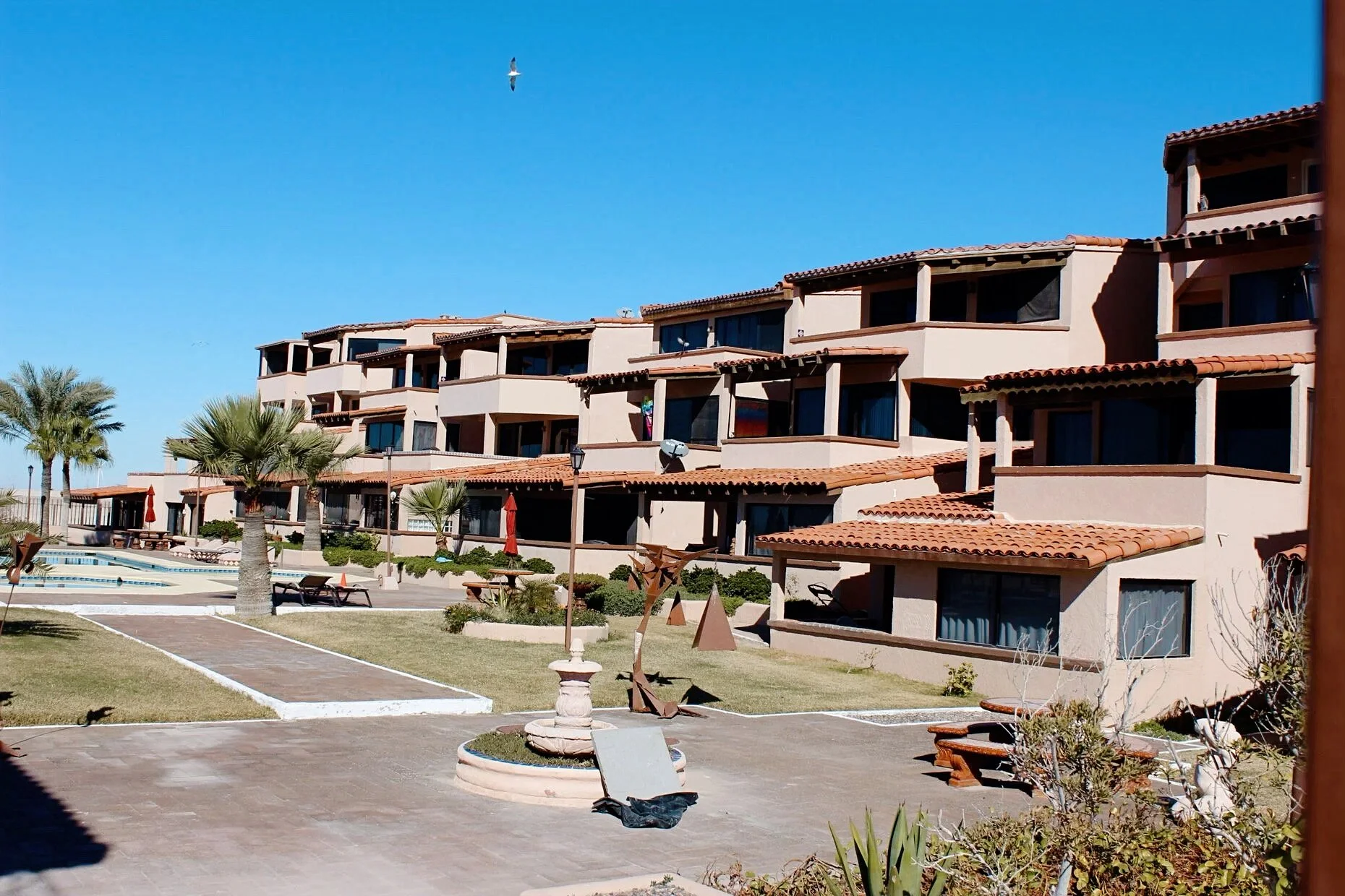 Mediterranean-style residential complex with beige buildings, red-tiled roofs, a pool, palm trees, and a clear blue sky.