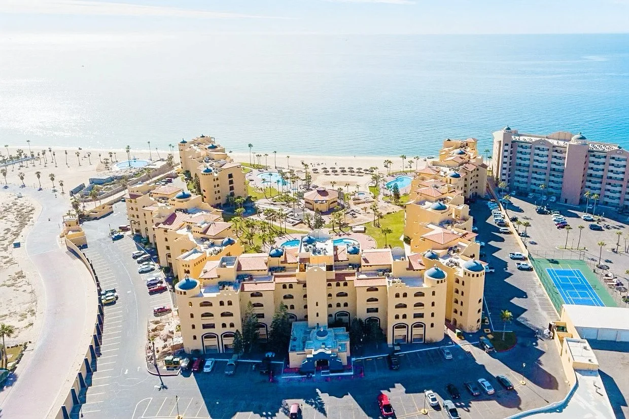 Aerial view of seaside resort with yellow buildings, multiple swimming pools, palm trees, and a beach overlooking the ocean.