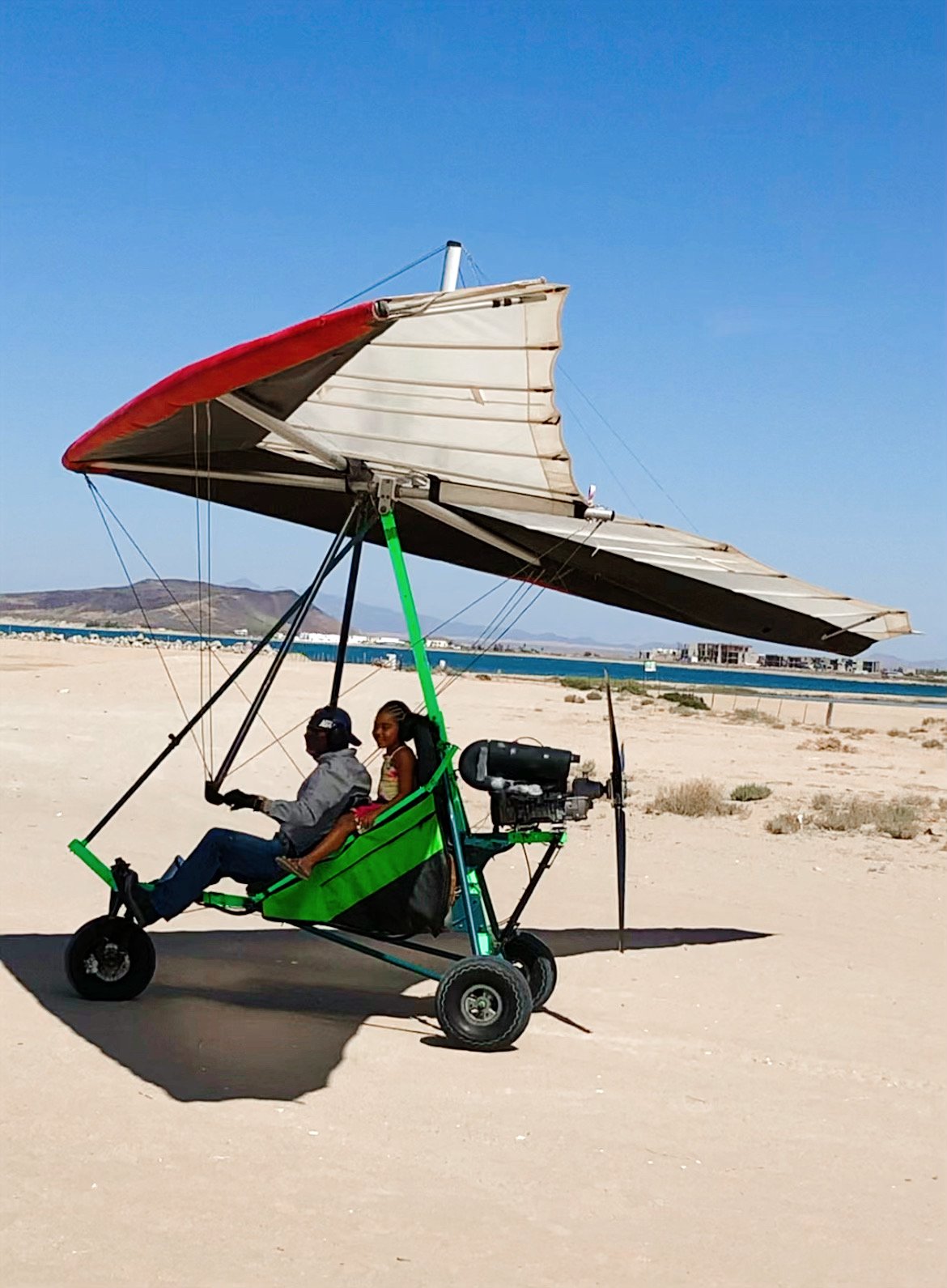 Two people sitting in a motorized hang glider on a sandy beach with a clear blue sky in the background.