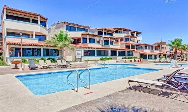 Outdoor pool area with lounge chairs and a multi-story building with balconies in the background, featuring Mediterranean-style architecture.