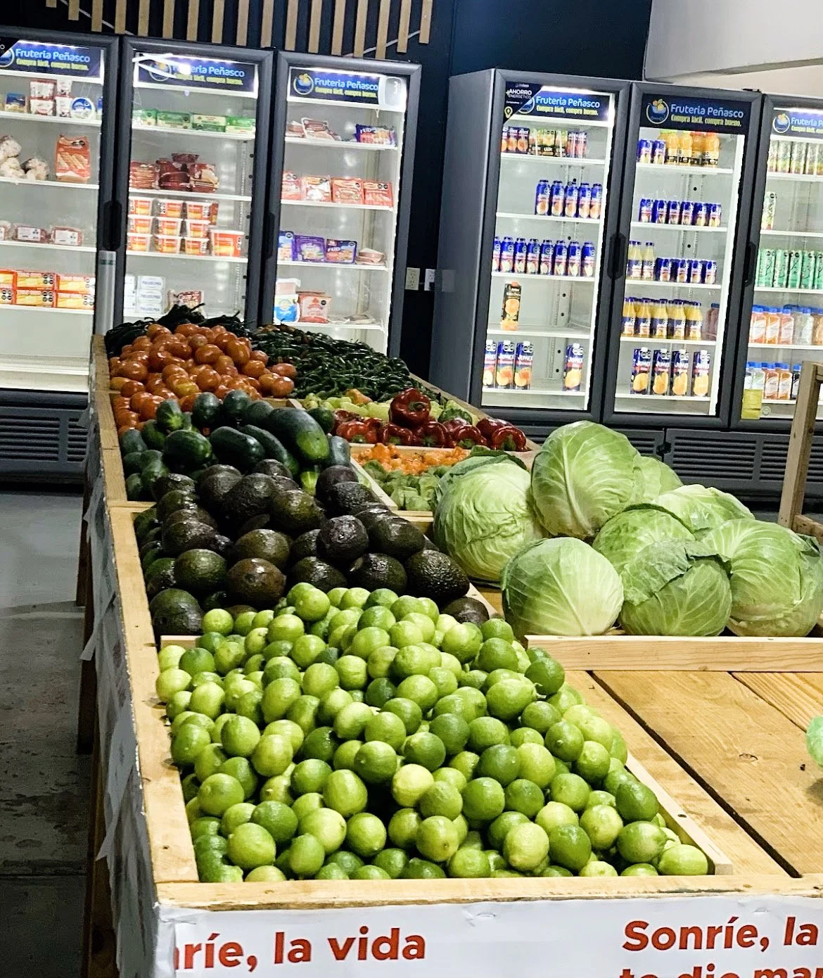 A supermarket produce section featuring piles of limes, avocados, and cabbages in the foreground. Refrigerated shelves with dairy and meat products are visible in the background.