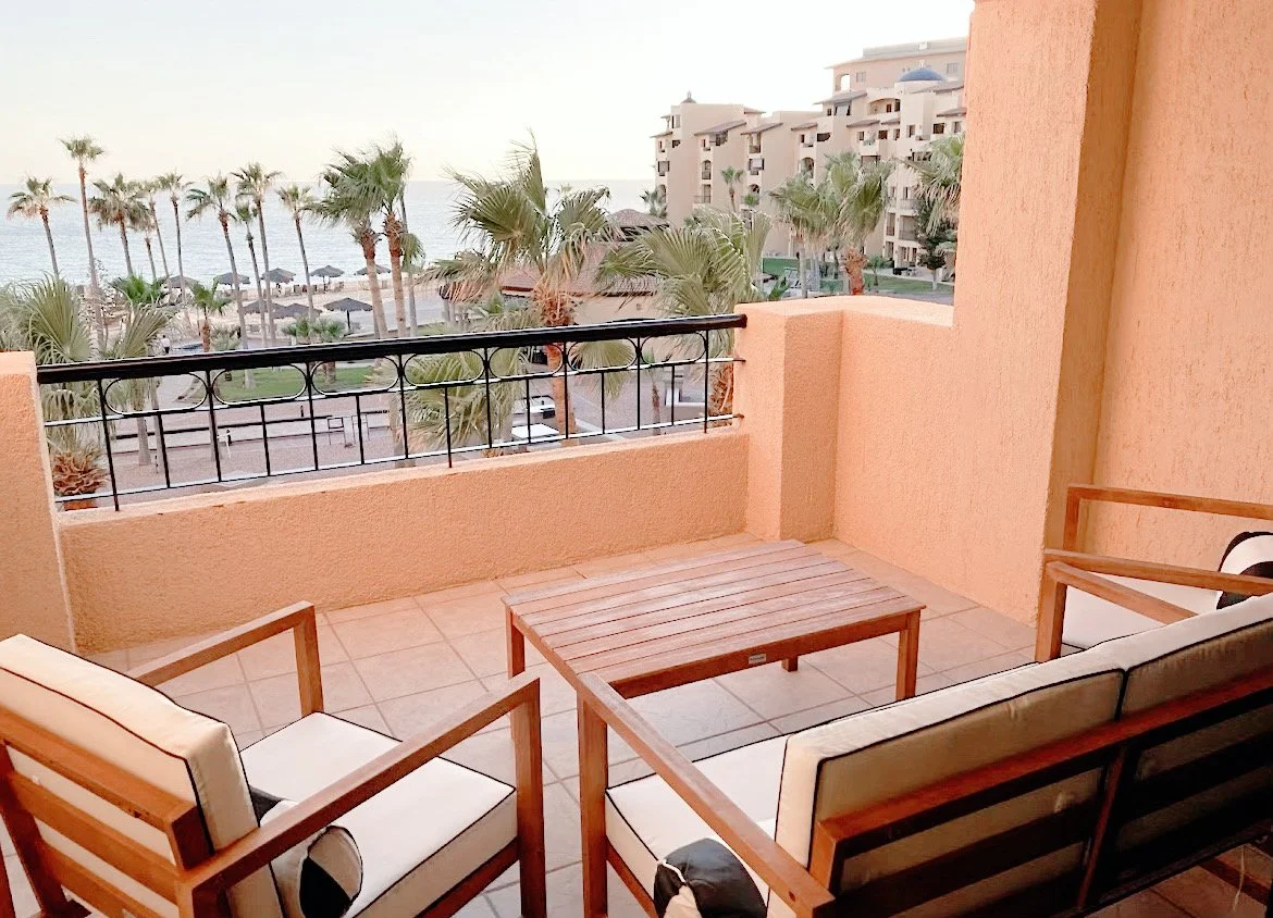 Balcony view with two cushioned chairs and a wooden table, overlooking palm trees, a beach, and the ocean at a resort.