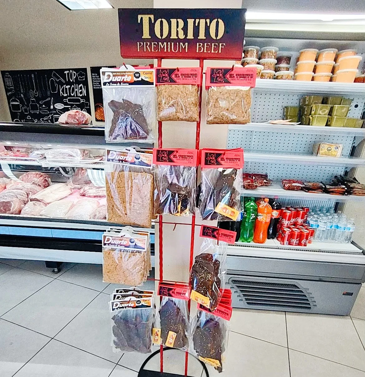 Display rack with Torito Premium Beef, dried meat packages, against a grocery store backdrop with a meat counter and refrigerator.