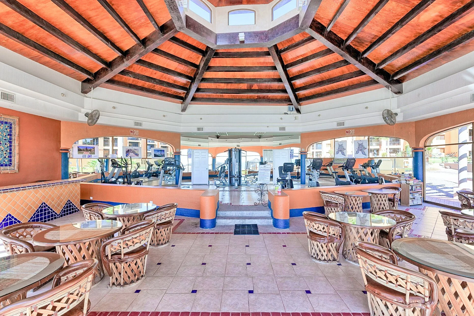 Indoor gym and seating area with glass tables and wicker chairs under a wooden ceiling, featuring exercise machines and decorative tiles on walls.