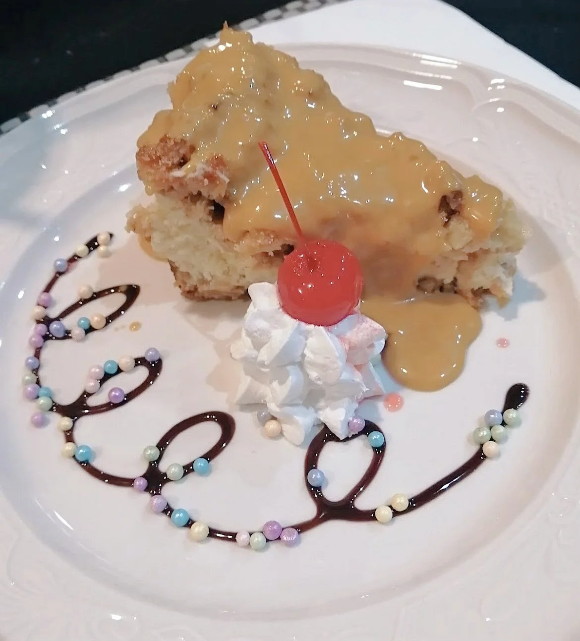 Cake slice with caramel sauce, whipped cream, maraschino cherry, chocolate drizzle, and candy pearls on a plate.