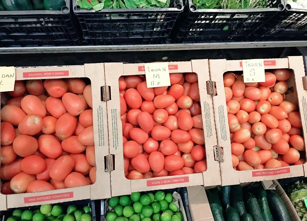 Boxes of vine-ripened tomatoes on display in a market, with limes and cucumbers below.