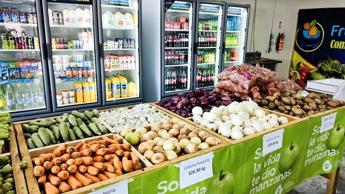 Grocery store produce section with fresh vegetables including carrots, onions, and cucumbers, alongside a refrigerated area with drinks and dairy products.
