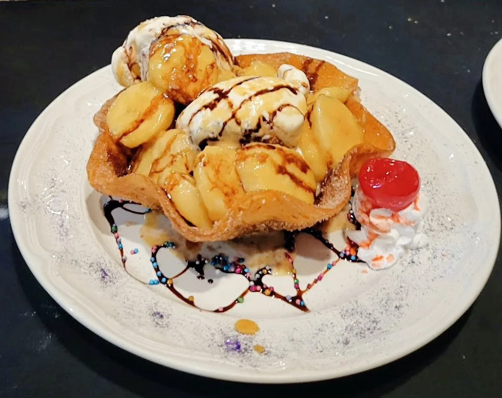 A dessert featuring a waffle bowl filled with vanilla ice cream, banana slices, caramel, and chocolate drizzle, topped with a maraschino cherry and whipped cream, with colorful sprinkles on the plate.