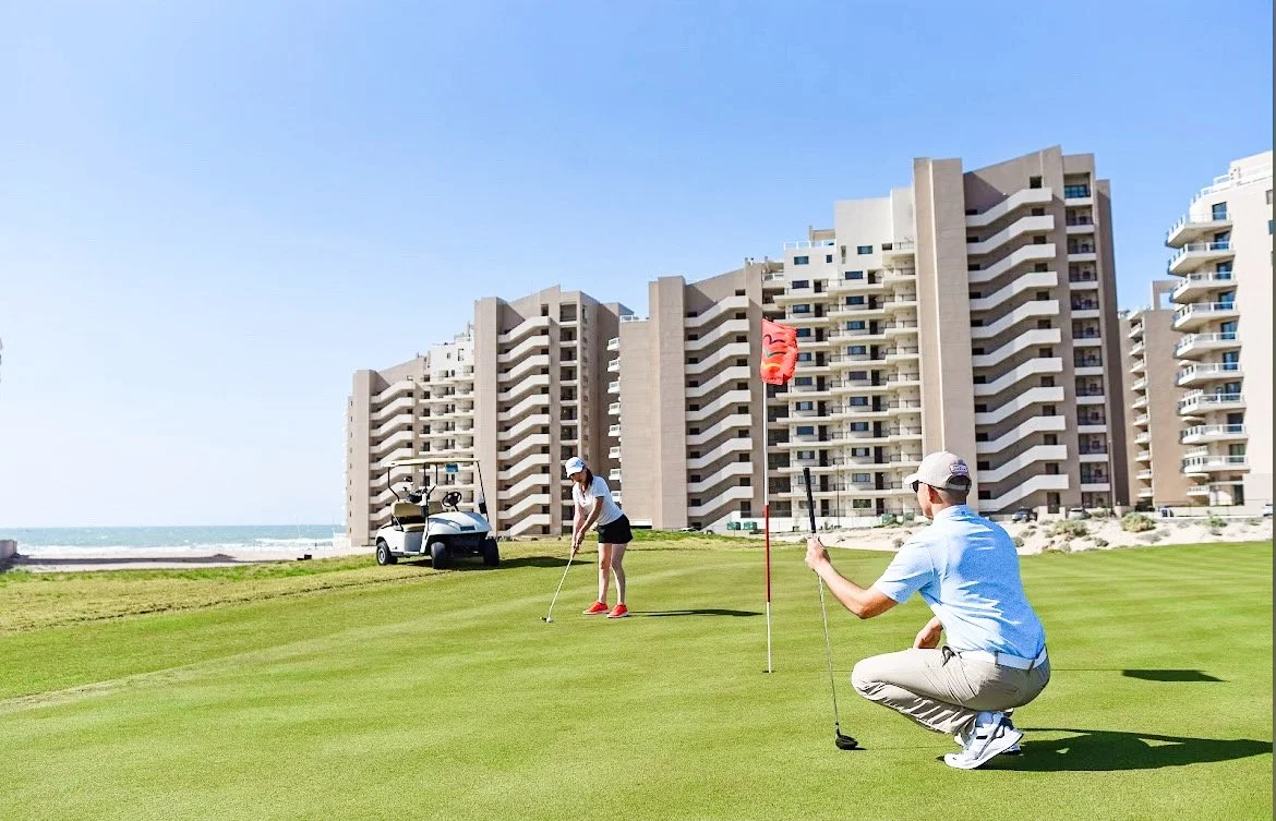Two golfers on a green near a flagstick with buildings in the background. A golf cart is parked nearby, and the ocean is visible in the distance under a clear blue sky.