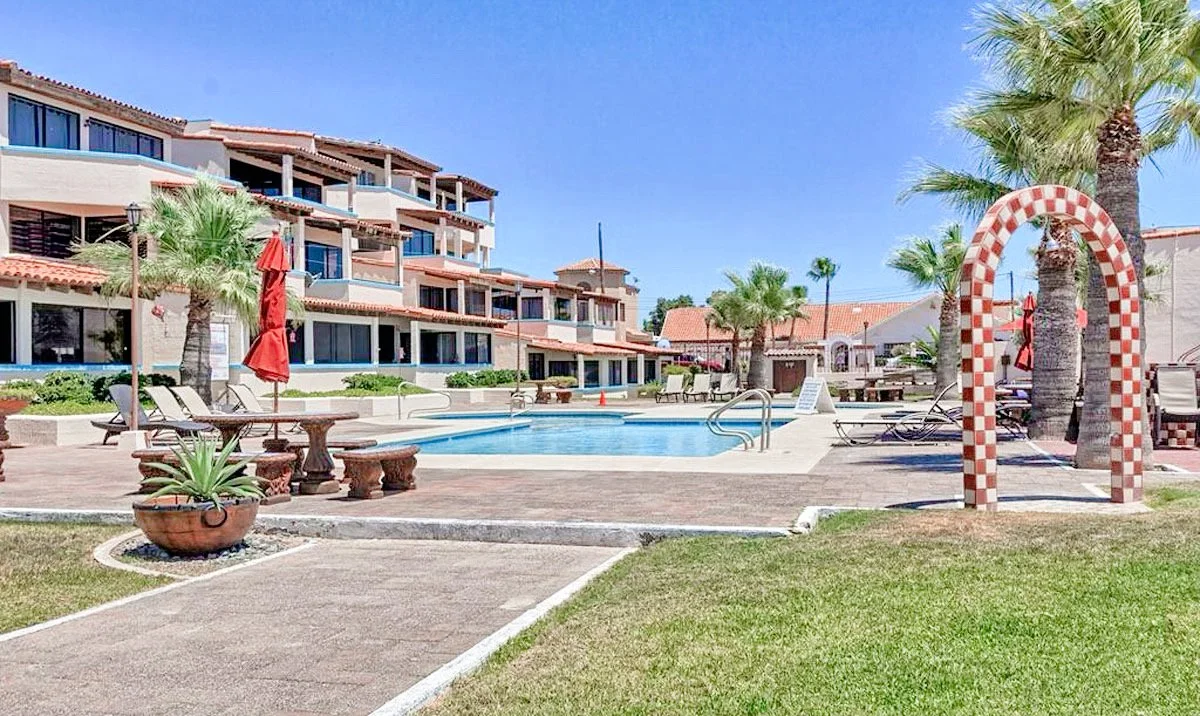 Resort outdoor pool area with palm trees, lounge chairs, red umbrellas, and arched tile feature.