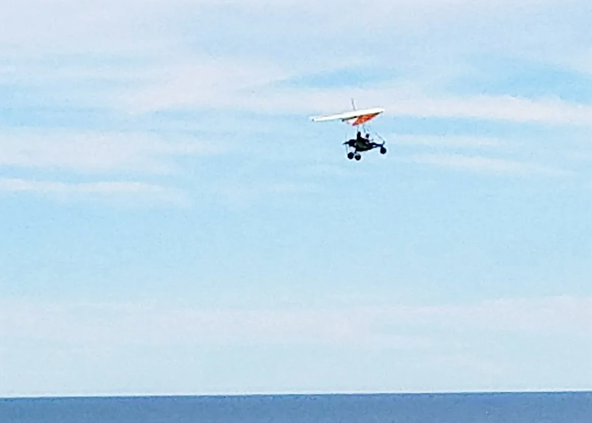 A microlight aircraft flying over the ocean with a blue sky backdrop.