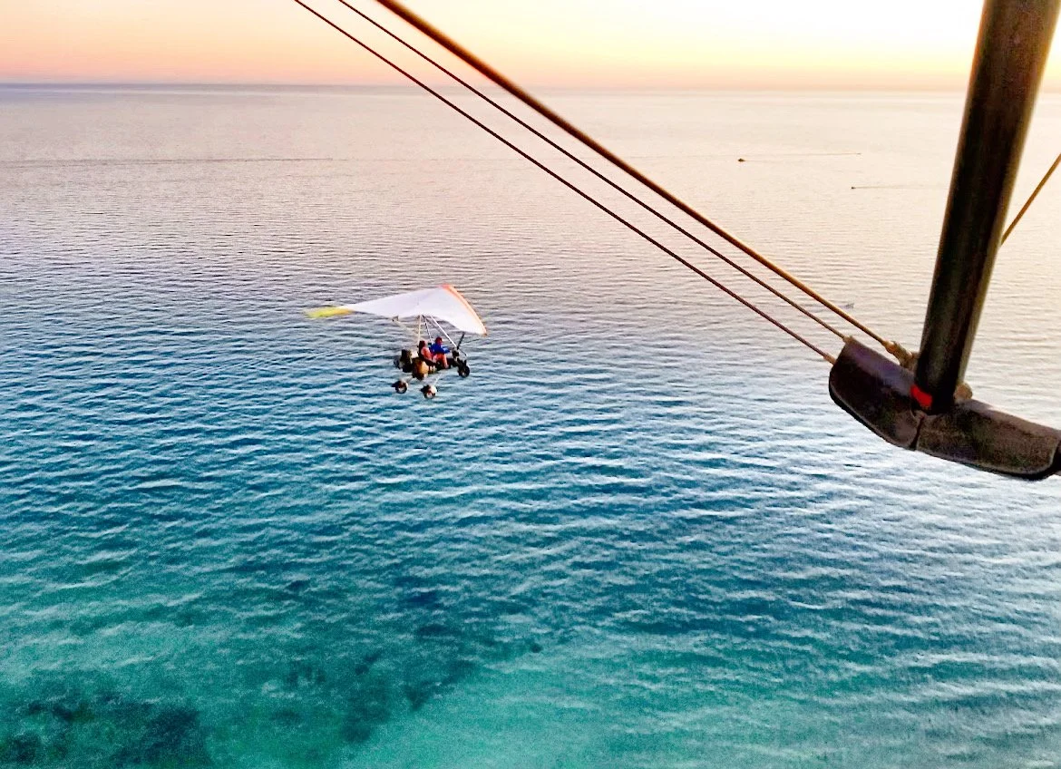 A powered hang glider flying over the ocean at sunset with clear skies.