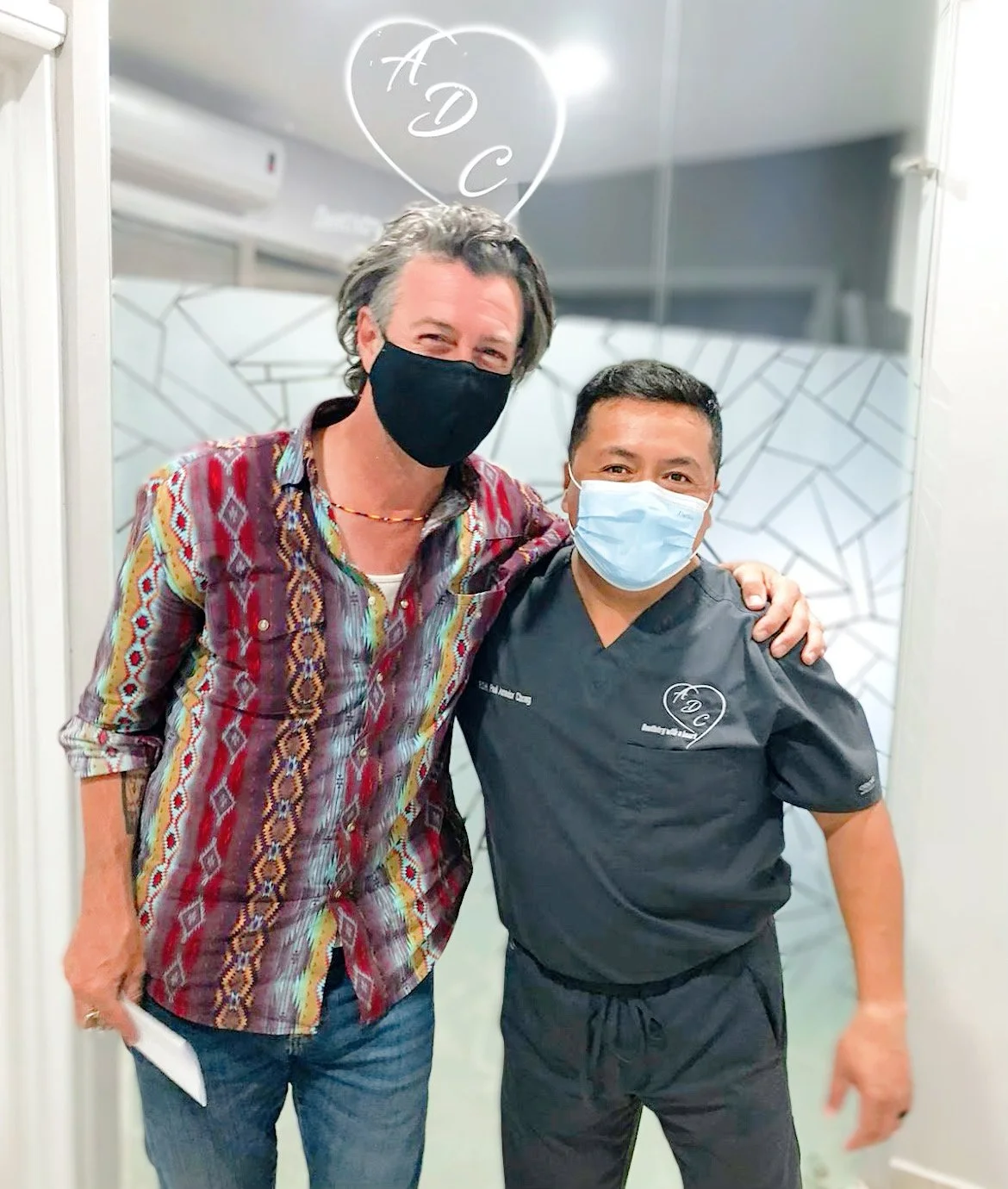 Two men wearing face masks, one in a colorful shirt and the other in black medical scrubs, standing together indoors.