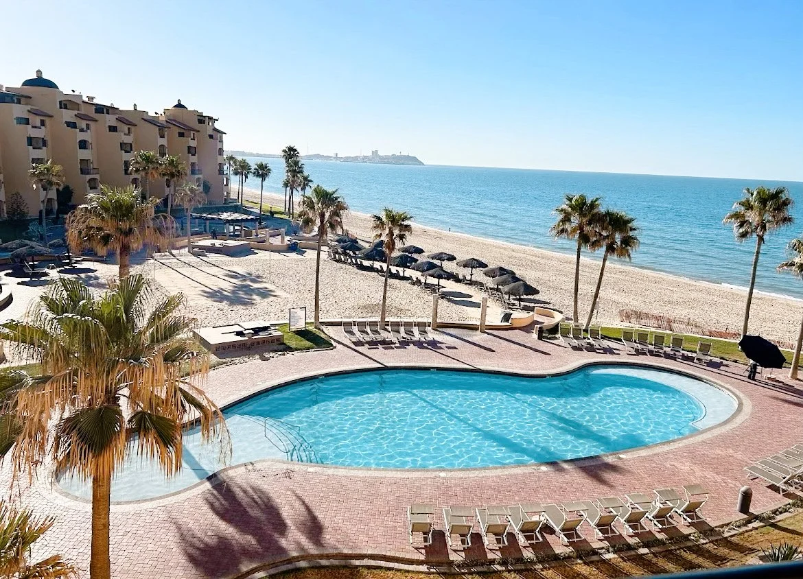 Beachfront resort with swimming pool, lounge chairs, palm trees, and ocean view under clear blue sky.