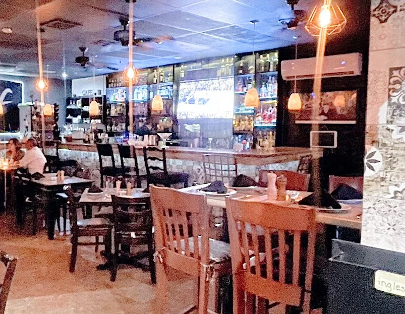 Interior of a dimly lit restaurant with a bar area, high chairs, and decorative hanging lights. Tables are set with black napkins and wooden chairs. Wall-mounted screens are visible in the background.