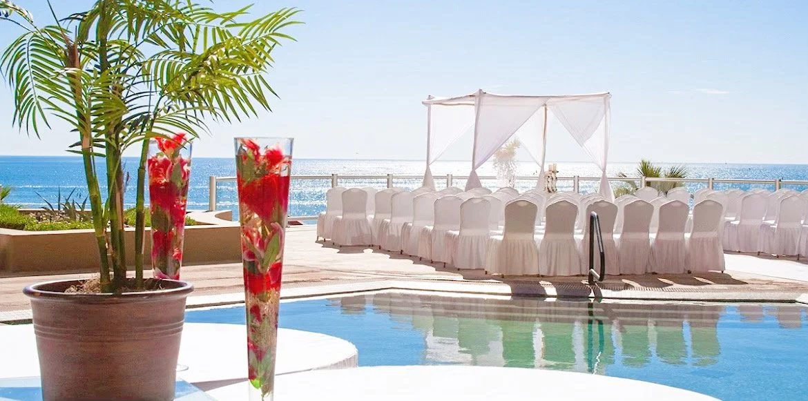 Outdoor wedding setup by a pool with white chairs and canopy, ocean view, and decorative red flowers in vases.