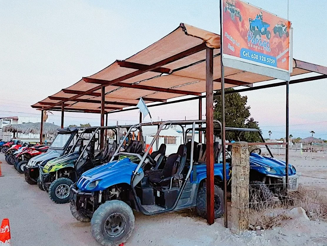Off-road vehicles parked under a canopy at a rental lot, identified by a sign overhead. The vehicles are side-by-sides, and the setting appears to be a desert area.