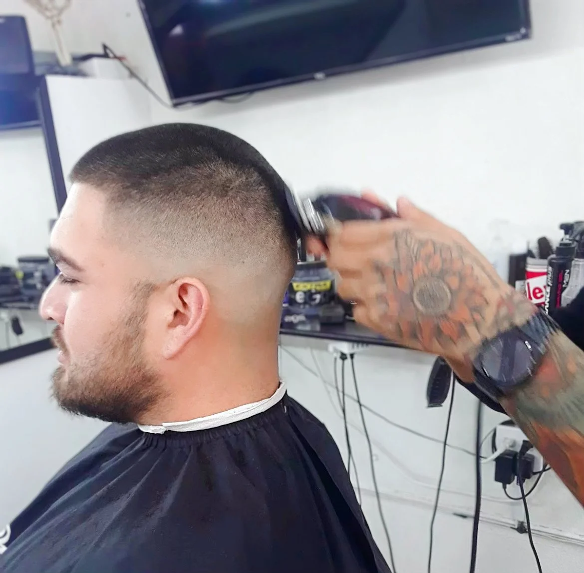 A person getting a haircut with clippers at a barbershop, performed by a barber with tattooed hands.