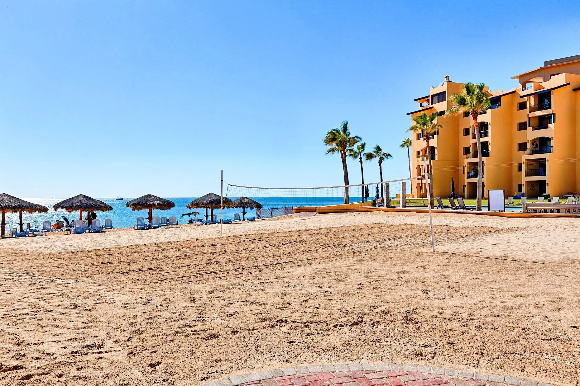 Beach scene with a volleyball court, straw umbrellas, lounge chairs, palm trees, ocean view, and a tan building.