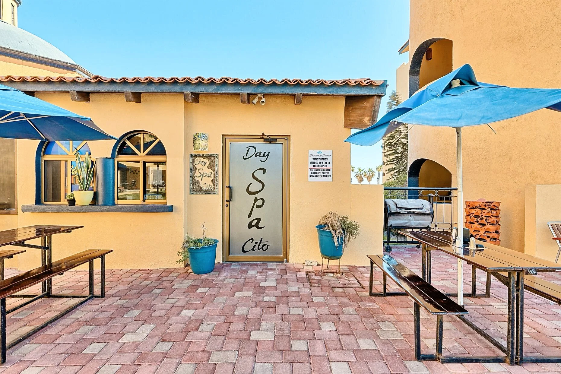 Entrance to a day spa in a sunny outdoor patio area with benches, tables, blue umbrellas, and potted plants.