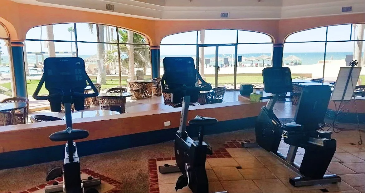 Indoor gym with exercise bikes facing large windows overlooking a beach view.