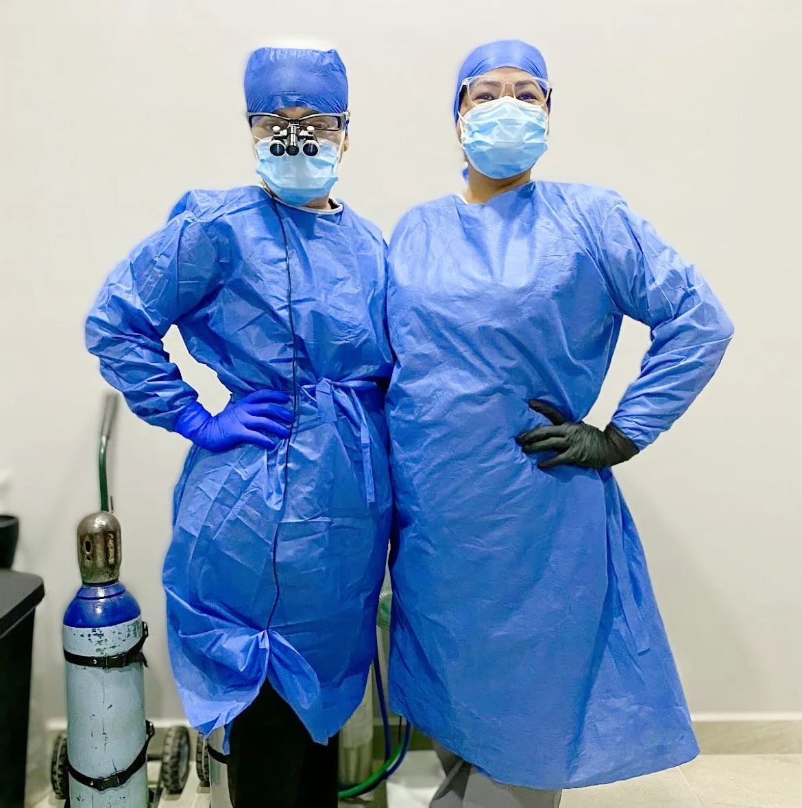 Two medical professionals wearing blue scrubs, masks, and gloves stand together. One wears magnifying glasses. An oxygen tank is in the background.