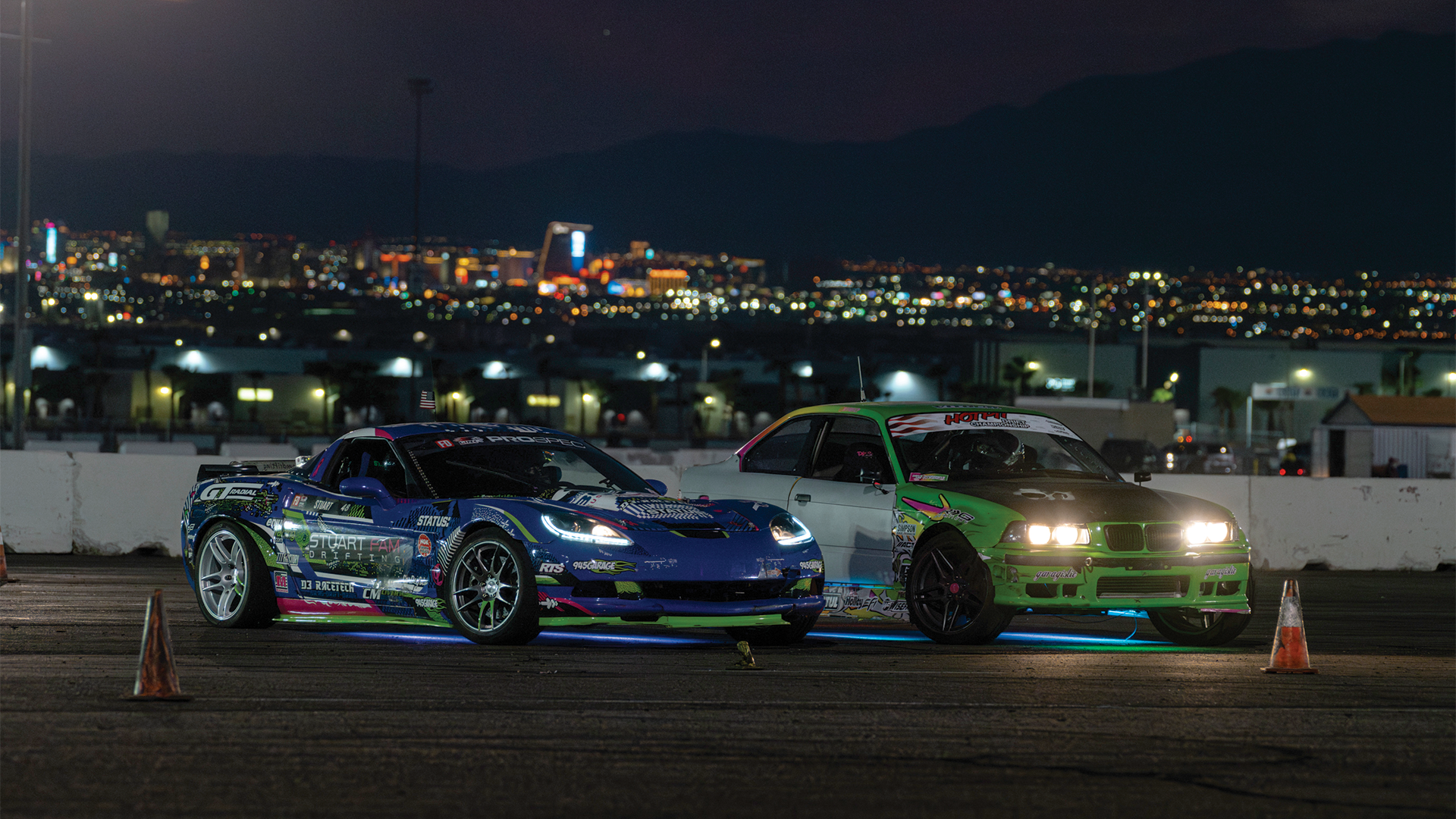 Two modified cars, a blue one on the left with racing decals and a green one on the right with headlights on, parked on a dirt surface at night with a city skyline and mountains in the background.